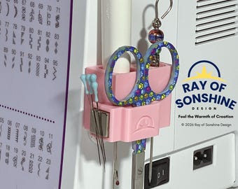 May include: A light pink plastic Magnetic Snip-n-Pick Sidecar organiser attached to a sewing machine. It holds a white tool, a pair of scissors with a colourful paw print pattern, and several sewing needles held by a square silver magnet.