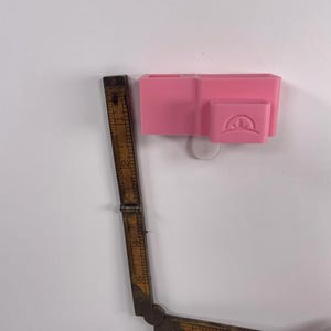 May include: A pink desk organizer with multiple compartments is mounted on a white wall. A vintage wooden folding ruler, with visible markings, is positioned next to the organizer. The ruler is partially extended, showing its length.