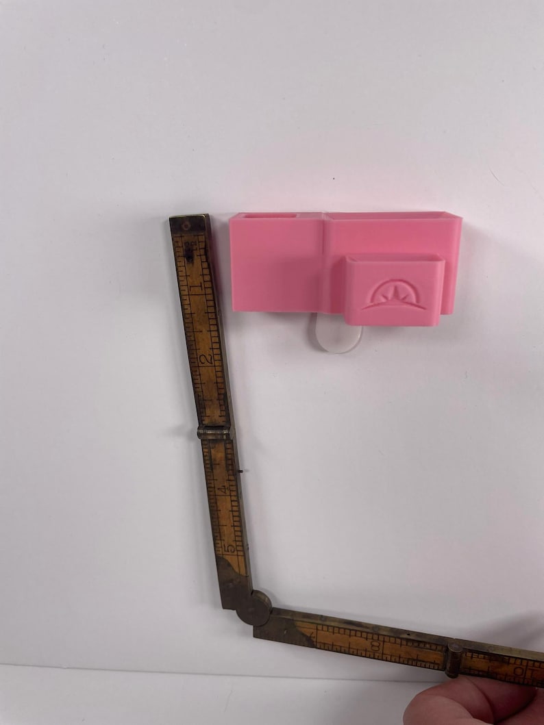 May include: A pink desk organizer with multiple compartments is mounted on a white wall. A vintage wooden folding ruler, with visible markings, is positioned next to the organizer. The ruler is partially extended, showing its length.