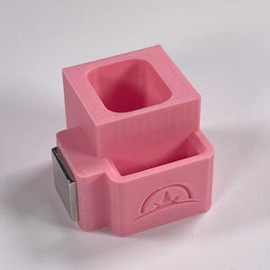 May include: A pink 3D-printed desk organizer with a rectangular top section and a lower section with a mountain design. A silver-colored magnet is attached to the side. The organizer is designed to hold small items.