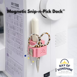 Puede incluir: Un soporte magnético rosa para tijeras y alfileres de costura, fijado a una máquina de coser blanca. Las tijeras tienen un diseño de estampado de leopardo. El texto "Magnetic Snip-n-Pick Dock" es visible.