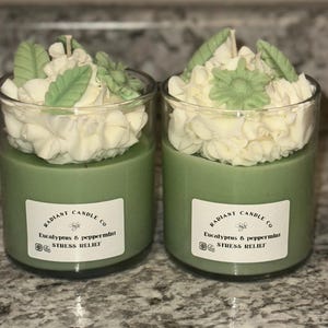 Peut inclure: Deux pots de bougies en verre vert avec un glaçage blanc et des décorations de feuilles vertes sur le dessus. Les étiquettes indiquent "RADIANT CANDLE CO Eucalyptus & peppermint STRESS RELIEF".