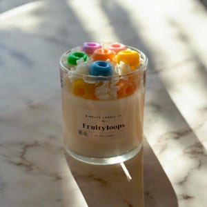 Puede incluir: Una vela de cera de soja blanca en un tarro de vidrio transparente con una etiqueta que dice "RADIANT CANDLE CO. Fruityloops". La vela está cubierta con bucles de cereales glaseados de colores.