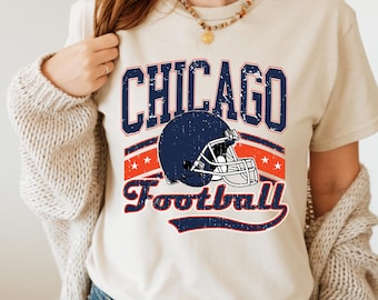 Könnte beinhalten: Cremefarbenes T-Shirt mit einem Used-Look-Footballhelm-Grafik in Marineblau und dem Text "CHICAGO Football" in Marineblau und Rot. Das Design enthält Sterne und Streifen und erinnert an eine Vintage-Sportästhetik.