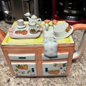May include: A whimsical ceramic teapot shaped like a kitchen stove, painted in peach and yellow. It features miniature teacups, a teapot, and a tray with strawberry designs. The teapot has a handle and spout.