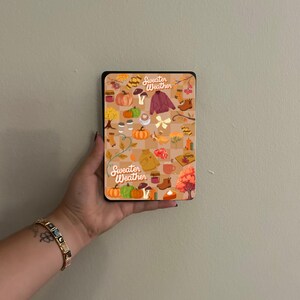 May include: A rectangular case with a black border featuring a fall-themed design. The design includes pumpkins, leaves, a sweater, boots, and the text "Sweater Weather" in a whimsical font. The case is held in a hand.
