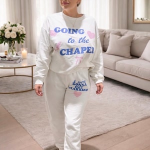 以下が含まれることがあります： 「GOING to the CHAPEL」の文字がプリントされた白いスウェットシャツと、「& we're GONNA GET MARRIED」の文字がプリントされた白いスウェットパンツのセットアップ。 文字は青とピンク。サンダルを履いています。