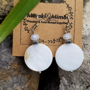 May include: A pair of drop earrings featuring large, circular white shell discs suspended from silver-toned metal hooks. Each earring includes a small, faceted iridescent bead and a metallic spacer bead above the shell disc. The earrings are displayed on a brown card with the text Meraki Mimos Passion & Soul thread together.