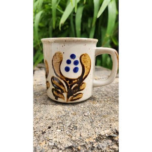May include: A ceramic mug with a beige speckled finish. The mug features a brown floral design with blue accents. The handle is a matching beige color. The mug is sitting on a stone surface.