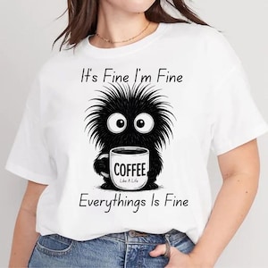 May include: White t-shirt featuring a black cartoon monster holding a coffee mug. The mug has the word "COFFEE" on it. The shirt also has the text "It's Fine I'm Fine, Everything's Is Fine."