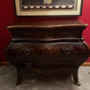 May include: A dark brown, antique-style chest of drawers with three drawers and ornate metal ring pulls. The chest has a curved design and decorative gold accents. A framed print with the text "MARCHE FLEURS" hangs above.