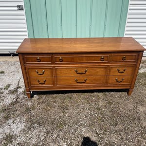 May include: A wooden dresser with a warm brown finish. It features nine drawers with brass-colored handles. The dresser has a rectangular shape and is set against a light green and white background.