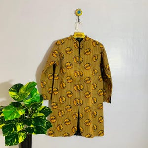 May include: A long, button-down coat in a mustard yellow color, adorned with a repeating pattern of yellow and brown circular designs. The coat has long sleeves and a mandarin collar. The garment is hanging on a hanger.