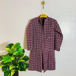 Puede incluir: Una chaqueta larga con botones de color burdeos oscuro con un diseño vertical estampado en blanco. La chaqueta está colgada en una percha amarilla contra una pared blanca. La chaqueta tiene mangas largas y cuello mandarín.