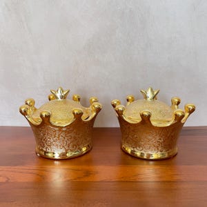 May include: Two gold-colored crown-shaped decorative containers with lids. The containers have a detailed floral pattern and are sitting on a wooden surface. Each crown has a small crown on top.