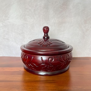May include: A round, lidded wooden bowl with a rich, dark red finish. The bowl features carved floral designs on the sides and lid, with a decorative knob on top. The bowl sits on a wooden surface.