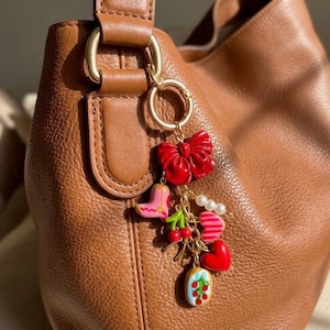 May include: A gold-toned keychain charm with a red bow, pink boot, cherries, hearts, and a small dessert. The charm is attached to a brown leather handbag with a gold ring and clasp.