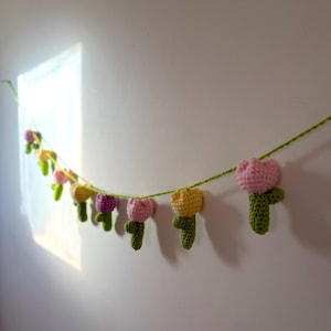 May include: A handmade crochet garland featuring a string of colorful tulips. The tulips are in shades of pink, yellow, and purple, with green stems and leaves. The garland is hanging against a white wall, creating a cheerful and decorative accent.