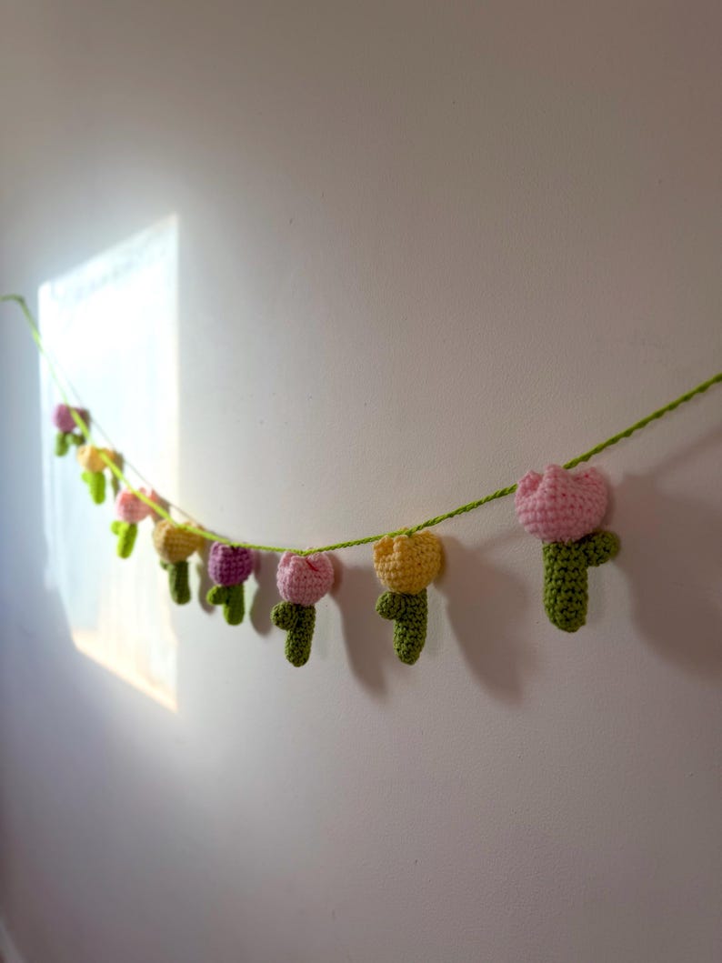 May include: A handmade crochet garland featuring a string of colorful tulips. The tulips are in shades of pink, yellow, and purple, with green stems and leaves. The garland is hanging against a white wall, creating a cheerful and decorative accent.