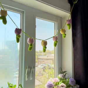 May include: A crocheted garland of colorful flowers hangs in front of a window. The garland features pink, yellow, and purple flowers with green stems. A basket of flowers and potted plants sit on the windowsill.