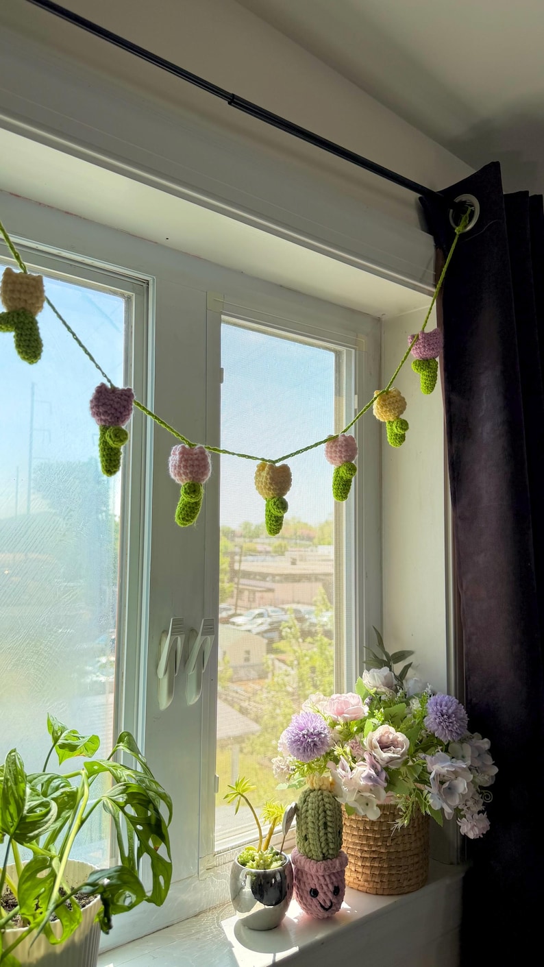 May include: A crocheted garland of colorful flowers hangs in front of a window. The garland features pink, yellow, and purple flowers with green stems. A basket of flowers and potted plants sit on the windowsill.