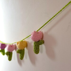 May include: A garland of crocheted tulips in various colors, including pink, yellow, and purple, strung on a green yarn. The handmade flowers are attached to a green stem, creating a decorative banner.