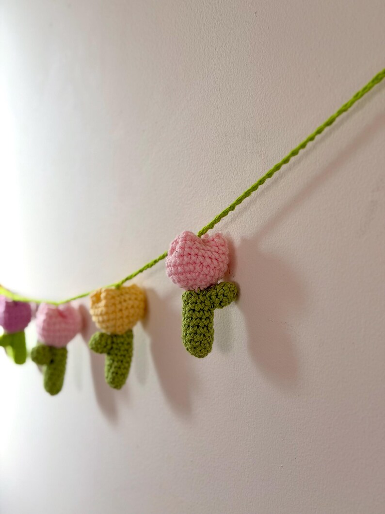 May include: A garland of crocheted tulips in various colors, including pink, yellow, and purple, strung on a green yarn. The handmade flowers are attached to a green stem, creating a decorative banner.