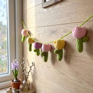 May include: A handmade crochet garland featuring colorful tulip flowers in pink, yellow, and purple. The flowers are strung on a green yarn and are attached to a light-colored wooden wall. The garland is a decorative item.