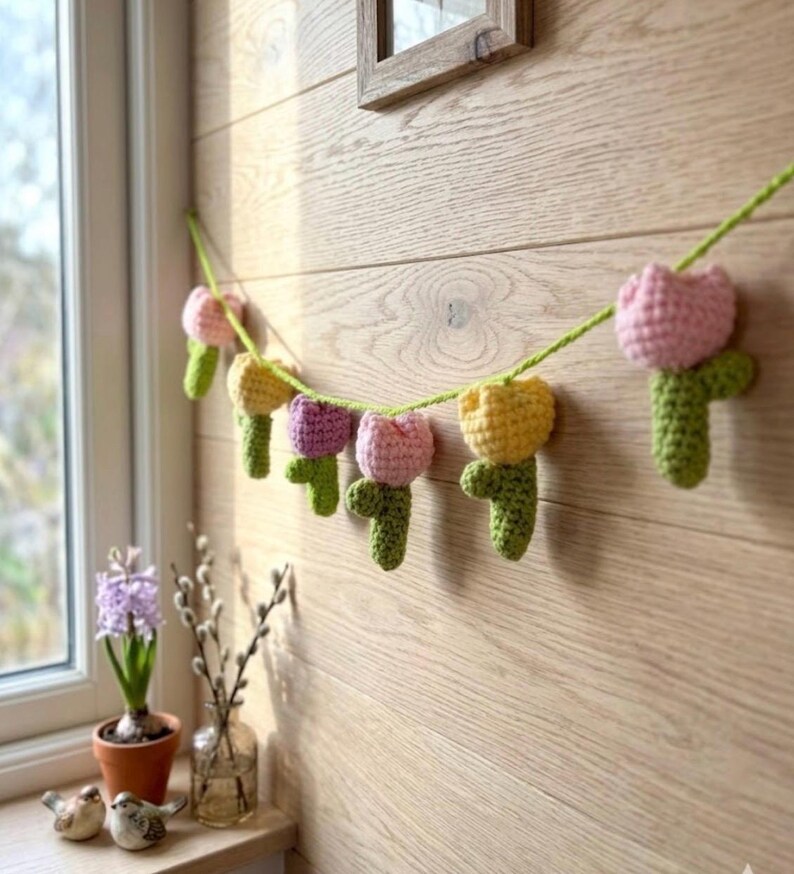 May include: A handmade crochet garland featuring colorful tulip flowers in pink, yellow, and purple. The flowers are strung on a green yarn and are attached to a light-colored wooden wall. The garland is a decorative item.