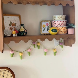 May include: A wooden shelf displays various decorative items. A framed cross-stitch, a floral vase, and ceramic bowls with fruit patterns are visible. A crocheted garland with pink and yellow flowers hangs below the shelf, adding a touch of handmade charm.