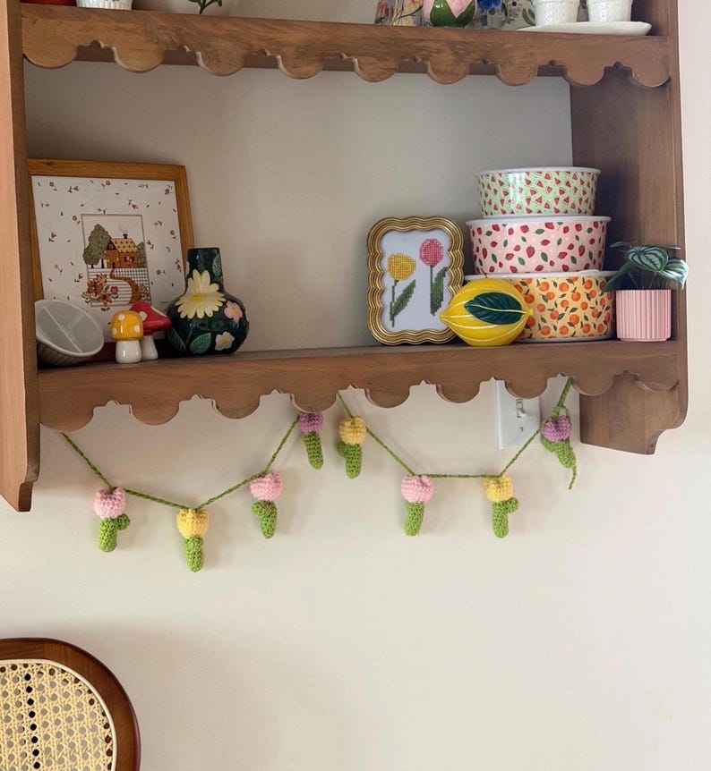 May include: A wooden shelf displays various decorative items. A framed cross-stitch, a floral vase, and ceramic bowls with fruit patterns are visible. A crocheted garland with pink and yellow flowers hangs below the shelf, adding a touch of handmade charm.