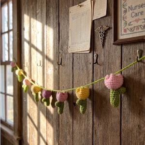 May include: A garland of crocheted tulips in shades of pink, yellow, and purple hangs against a rustic wooden wall. The handmade flowers are strung on a green yarn, creating a charming, spring-themed decoration.