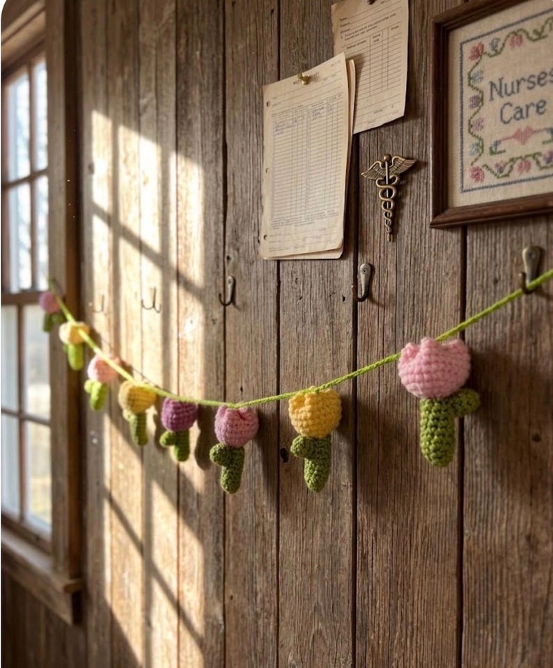 May include: A garland of crocheted tulips in shades of pink, yellow, and purple hangs against a rustic wooden wall. The handmade flowers are strung on a green yarn, creating a charming, spring-themed decoration.