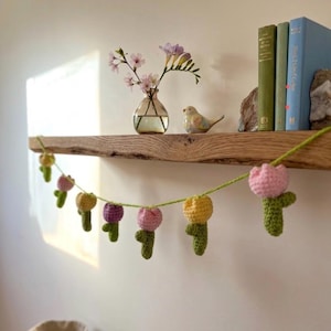 May include: A handmade crochet garland featuring colorful tulip flowers in pink, yellow, and purple. The garland is strung on green yarn and is displayed on a wooden shelf with books, a vase of flowers, and a decorative bird.