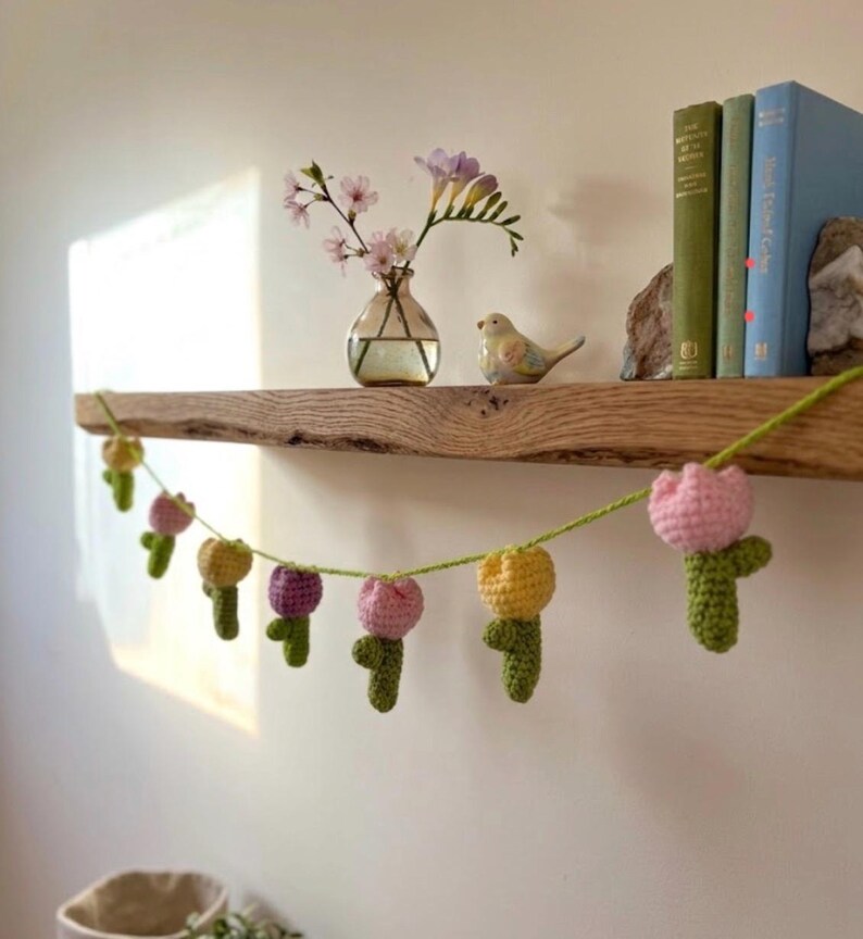 May include: A handmade crochet garland featuring colorful tulip flowers in pink, yellow, and purple. The garland is strung on green yarn and is displayed on a wooden shelf with books, a vase of flowers, and a decorative bird.