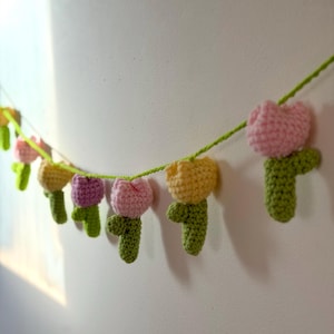May include: A crochet garland featuring multiple colorful tulip flowers. The tulips are in shades of pink, yellow, and purple, with green stems and leaves. The garland is strung on a green yarn and is hanging against a white wall.