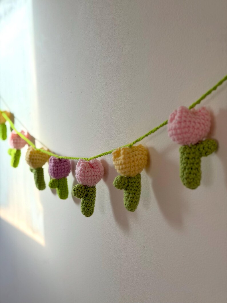 May include: A crochet garland featuring multiple colorful tulip flowers. The tulips are in shades of pink, yellow, and purple, with green stems and leaves. The garland is strung on a green yarn and is hanging against a white wall.