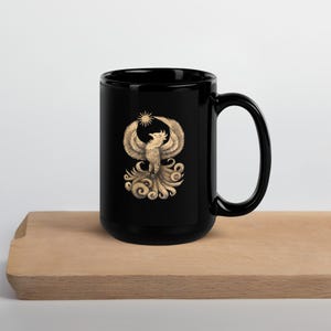 May include: Black ceramic mug featuring a detailed illustration of a phoenix with outstretched wings, a sunburst above its head, and swirling details below. The design is in shades of gold and tan. The mug has a large handle and is sitting on a wooden surface.