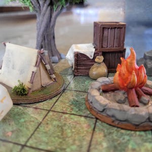 May include: A miniature tabletop gaming set featuring a beige tent, a campfire with orange and red flames surrounded by grey stones, two brown wooden crates, a small tan sack, a grey tree trunk, and a white twenty-sided die with gold numbers.