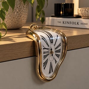 May include: A gold-toned melting clock with a white face and black Roman numerals. The clock features a curved, distorted frame designed to drape over the edge of a flat surface, with black hour and minute hands centred on the dial.