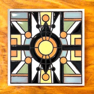 May include: A square ceramic tile with a geometric design in black, white, yellow, orange, and blue. The design features a central circle with a smaller circle inside, surrounded by radiating lines and geometric shapes.