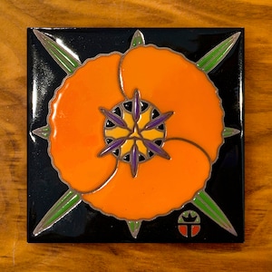 May include: A square ceramic tile with a black background and a stylized orange poppy flower with a purple and yellow center. The tile has a green and silver border and a small logo in the bottom right corner.
