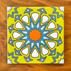 May include: A yellow ceramic tile with a blue, white, and orange geometric pattern. The pattern is a starburst design with a central orange star.
