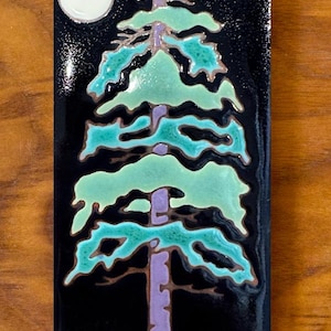 May include: A ceramic tile with a stylized image of a pine tree with a purple trunk and green branches against a black background. A white moon is visible in the upper left corner of the tile.