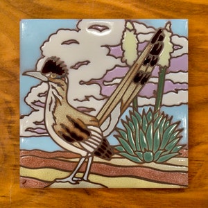 May include: A ceramic tile with a painted image of a roadrunner bird standing in a desert landscape. The bird is brown and white with a long tail. The background is blue with white clouds and a purple sky. There is a green agave plant in the foreground.