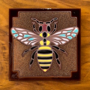 May include: A ceramic tile with a brown background and a detailed illustration of a bee with yellow and black stripes, blue and white wings, and red accents.