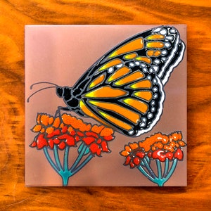 May include: A ceramic tile with a painted image of a monarch butterfly with orange, black, and white wings, perched on a cluster of red and orange flowers with green stems.