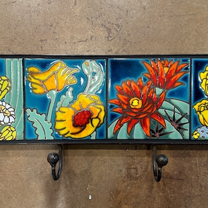 May include: A ceramic wall-mounted key holder with four hooks. The holder is decorated with four colorful tiles depicting stylized desert flowers and cacti. The tiles are in shades of blue, green, yellow, orange, and red.