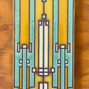 May include: A rectangular ceramic tile with a geometric design in shades of yellow, green, and white. The design features vertical lines and squares, creating a symmetrical pattern.