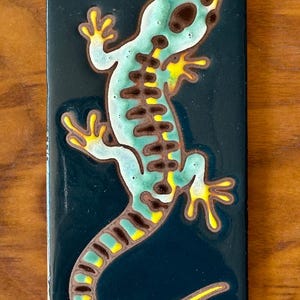 May include: A ceramic tile with a blue background and a yellow, green, and brown lizard design.